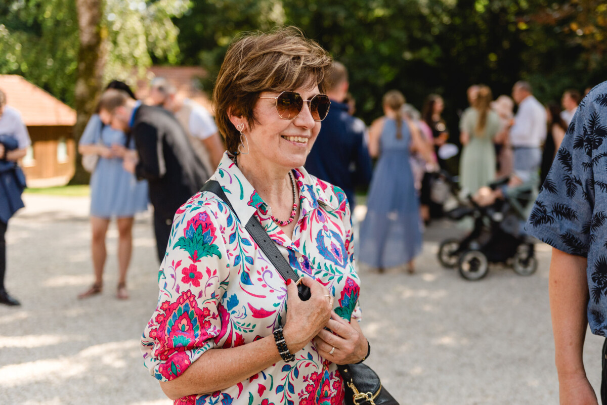 Eine Frau mit Sonnenbrille und geblümter Bluse steht im Freien und hält eine Handtasche in der Hand, im Hintergrund unterhalten sich Leute.