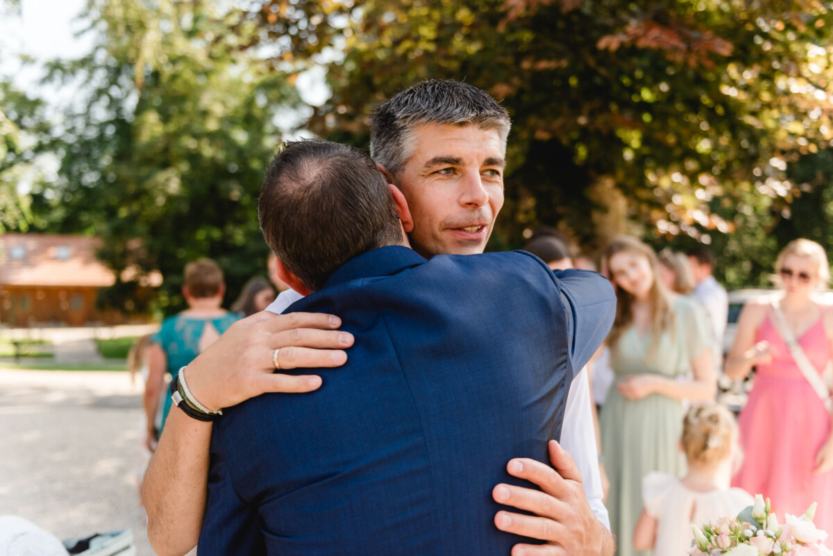 Zwei Männer umarmen sich bei einem geselligen Beisammensein im Freien. Die Menschen im Hintergrund sind unter Bäumen in legerer und formeller Kleidung gekleidet.