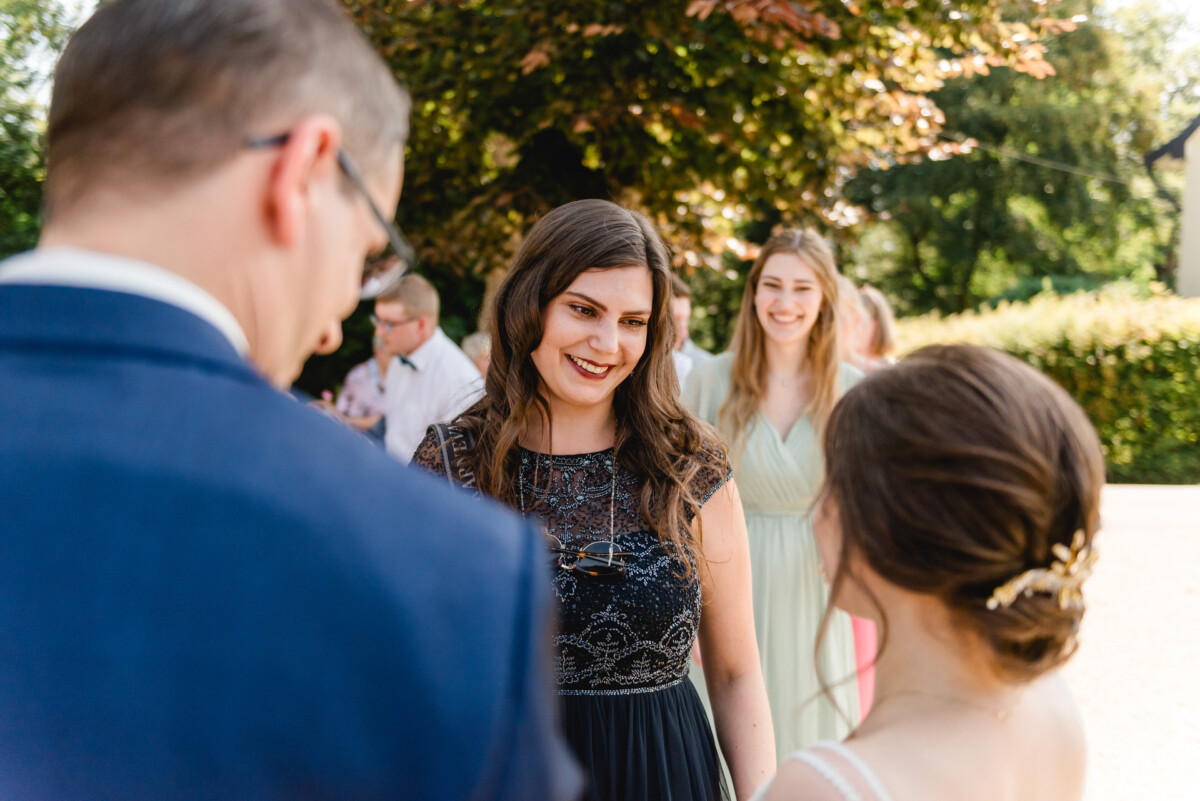 Eine Gruppe von Menschen steht draußen, lächelt und unterhält sich. In der Mitte steht eine Frau in einem schwarzen Kleid. Im Hintergrund sind Bäume zu sehen.