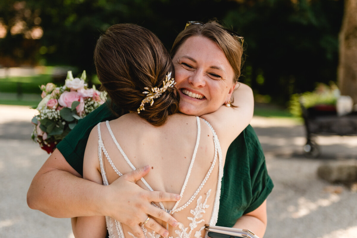 Zwei Frauen umarmen sich herzlich im Freien; eine hält einen Blumenstrauß und die andere lächelt breit.
