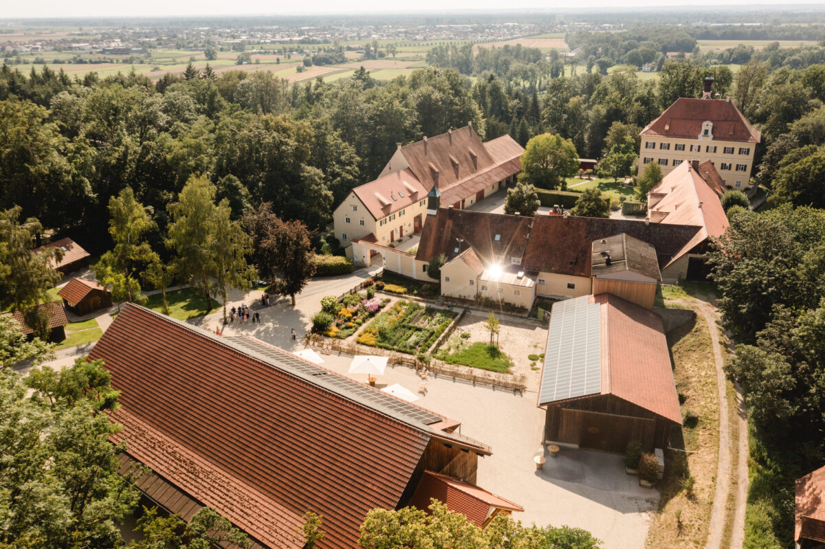 Luftaufnahme eines großen Anwesens mit mehreren Gebäuden mit roten Dächern, umgeben von Bäumen und offenen Feldern unter einem klaren Himmel.