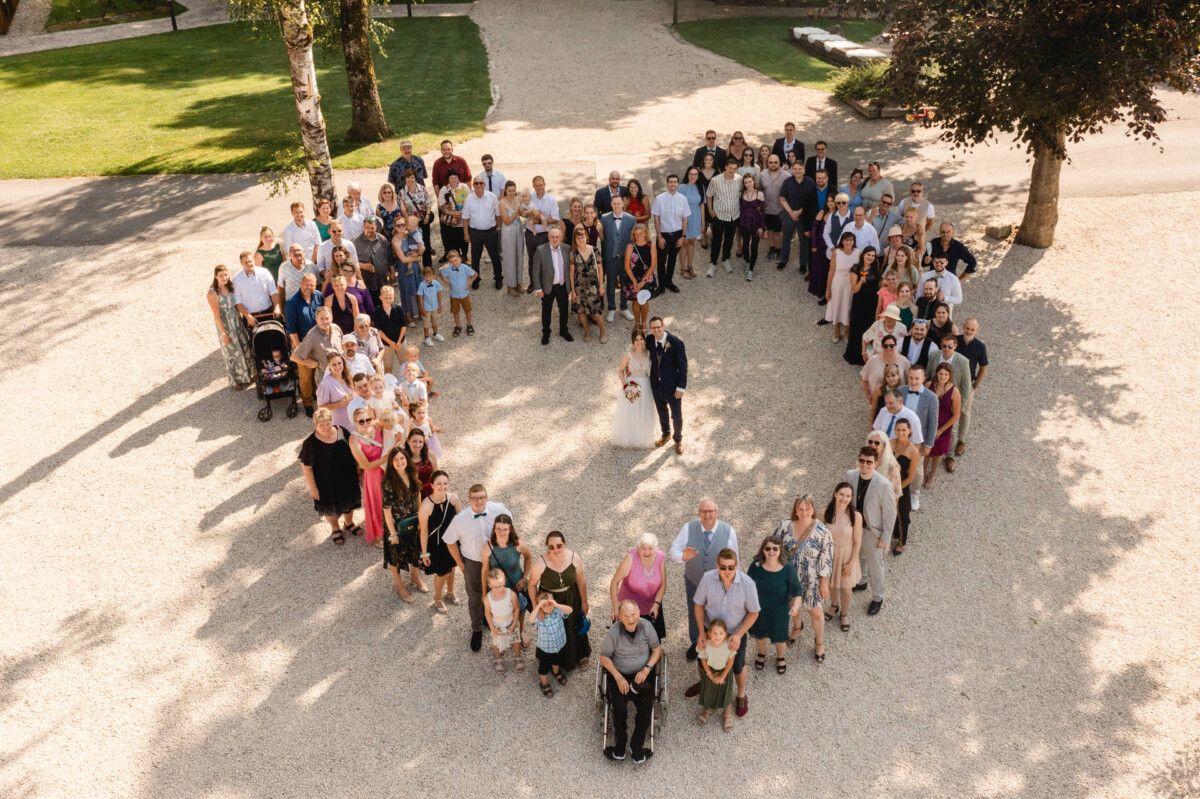 Eine große Gruppe von Menschen, die in Form eines Herzens angeordnet sind, mit einem Paar in der Mitte. Die Szene ist im Freien auf einer Kiesfläche mit umgebendem Grün.