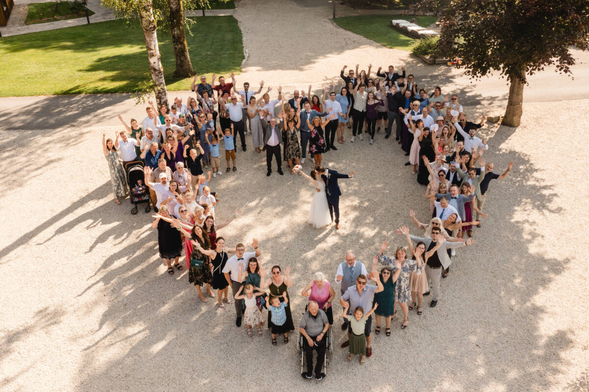 2024.07.27-Beatrice-Christian-Gut-Mergenthau-Kissing_01004 Eine große Gruppe von Menschen, die im Freien in Herzform angeordnet sind, wobei einige ihre Hände heben. Im Hintergrund sind Bäume und ein Weg zu sehen.
