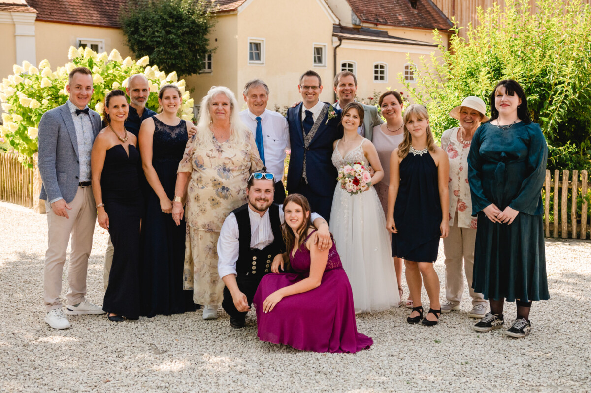 Eine Gruppe von Menschen, darunter Braut und Bräutigam, posiert im Freien vor einem Haus. Einige sind formell gekleidet, andere leger. Im Hintergrund sind Blumen und Bäume zu sehen.