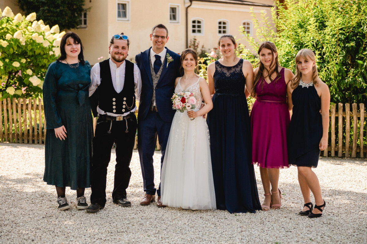 Eine Braut und ein Bräutigam stehen mit fünf Personen draußen. Die Gruppe ist halbformell gekleidet, einige tragen Kleider und eine Weste und Hose. Im Hintergrund sind ein Holzzaun und Grünpflanzen zu sehen.