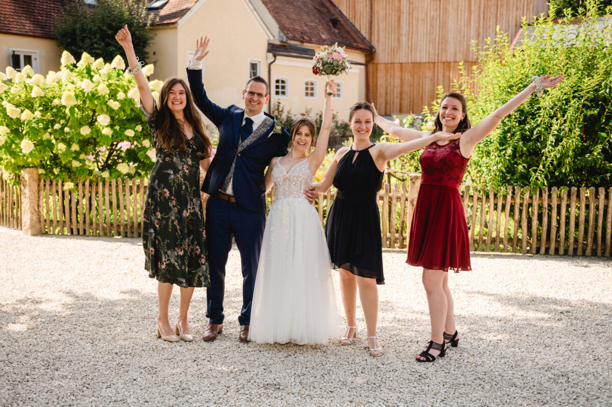 Eine Braut und ein Bräutigam stehen mit drei anderen Personen im Freien, alle lächeln und heben die Arme. Sie stehen vor einem Holzzaun und grünen Sträuchern und tragen formelle Kleidung.