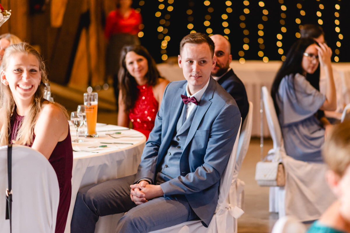 Ein Mann in einem blauen Anzug und einer Fliege sitzt mit anderen Teilnehmern einer formellen Veranstaltung an einem Tisch.
