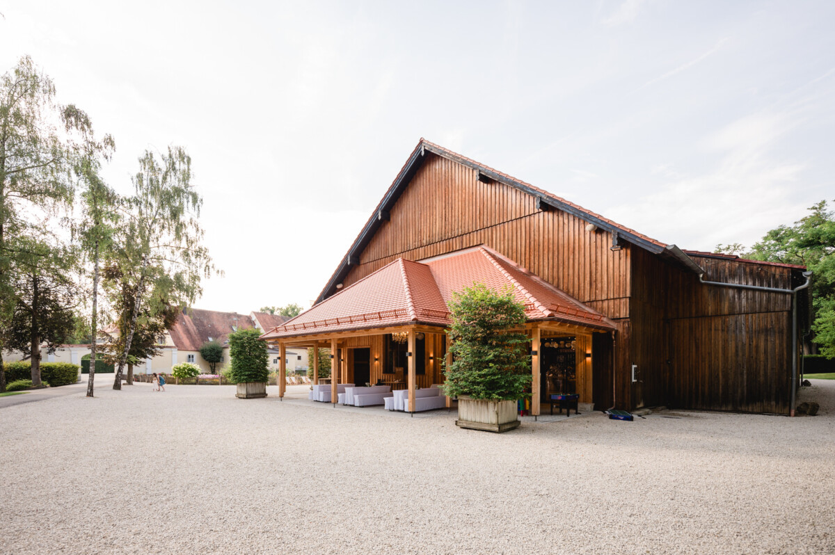 Eine große Holzscheune mit rotem Dach, umgeben von Bäumen und einem Kieshof an einem klaren Tag.
