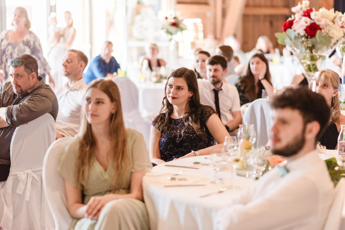 Gäste sitzen an Tischen und verfolgen aufmerksam ein Geschehen in einem dekorierten Veranstaltungsort.