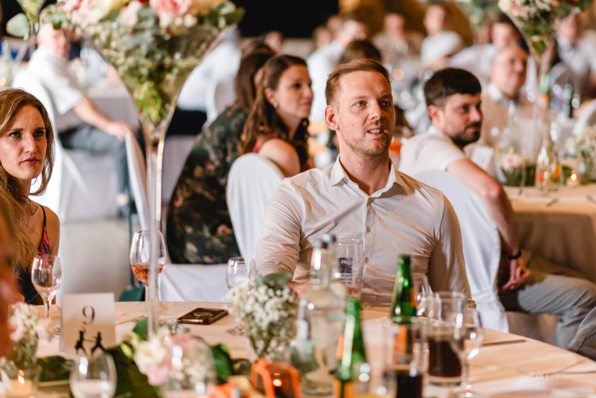 Menschen sitzen an Tischen mit Blumenschmuck und hören bei einer Veranstaltung aufmerksam zu.