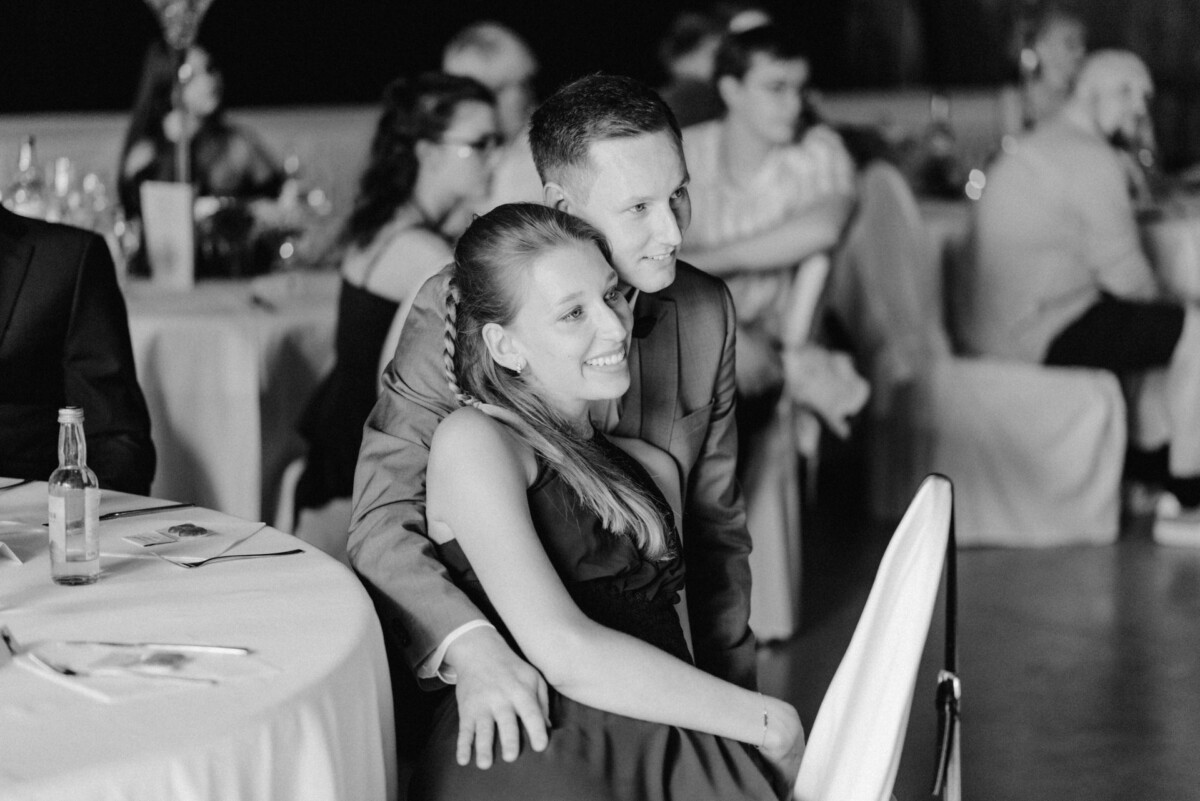 Bei einer formellen Veranstaltung sitzt ein Paar eng beieinander, umgeben von den anderen Gästen an den Tischen. Die Atmosphäre wirkt entspannt und gesellig.