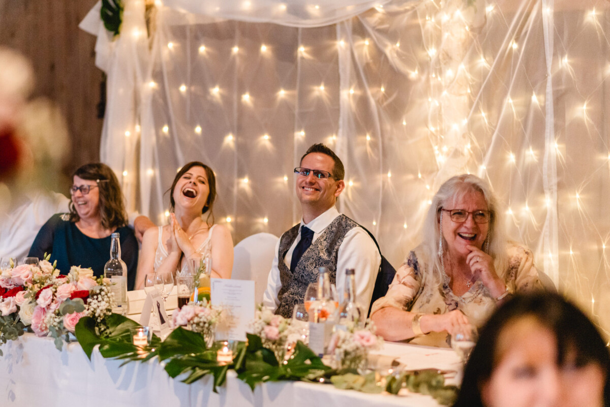 Menschen sitzen an einem dekorierten Tisch, lächeln und lachen, mit Blumenarrangements und funkelnden Lichtern im Hintergrund.