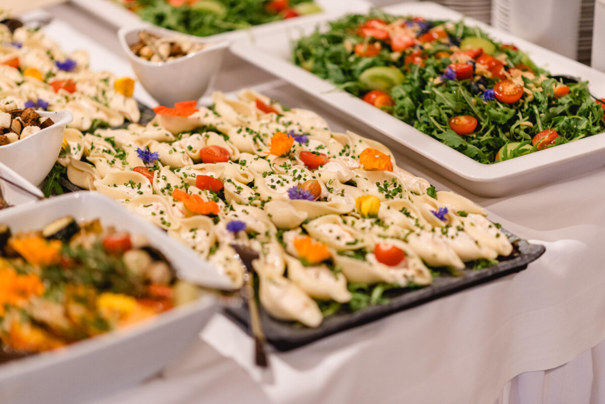 Ein Buffettisch mit verschiedenen Gerichten, darunter mit Kräutern und Blumen garnierte Knödel, ein Salat mit Kirschtomaten und geröstetes Gemüse.