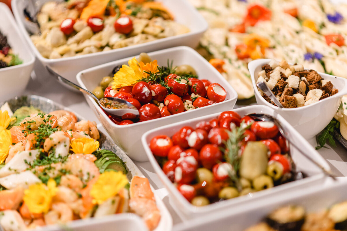 Auf einem Tisch werden verschiedene Tellergerichte mit Kirschtomaten, Oliven, Garnelen und garniert mit Kräutern und essbaren Blumen präsentiert.