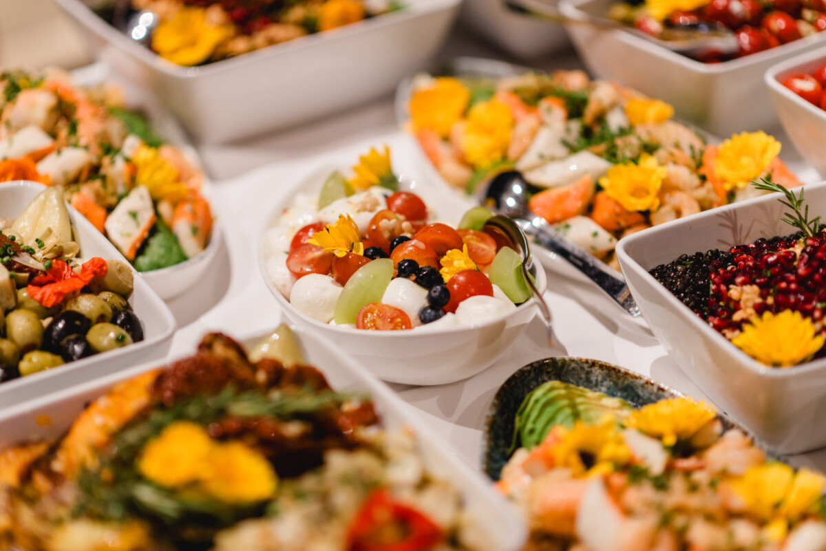 Eine Auswahl bunter Salate mit verschiedenen Früchten, Gemüsen und Blumen in weißen Schüsseln auf einem Tisch.