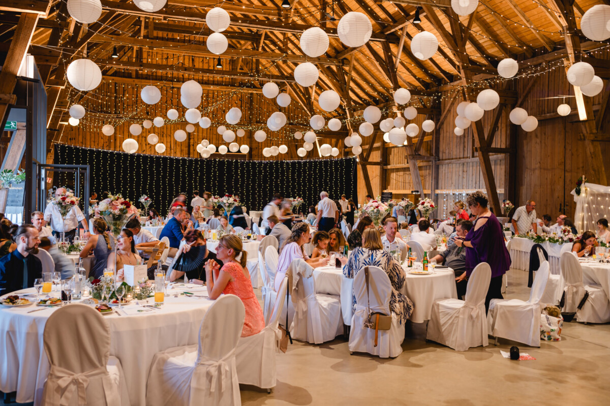 Eine große Versammlung in einem dekorierten Holzsaal, bei der die Gäste an runden, mit weißen Tüchern bedeckten Tischen sitzen. Hängende Papierlaternen und Lichterketten sorgen für eine festliche Atmosphäre.