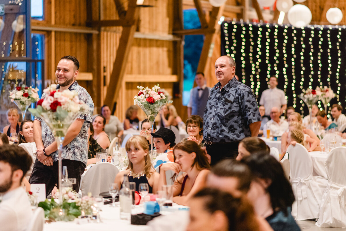 Eine Gruppe von Menschen sitzt an Tischen in einem mit Lichterketten und Blumen geschmückten Holzsaal. Zwei Männer stehen da und schauen sich eine Veranstaltung an.
