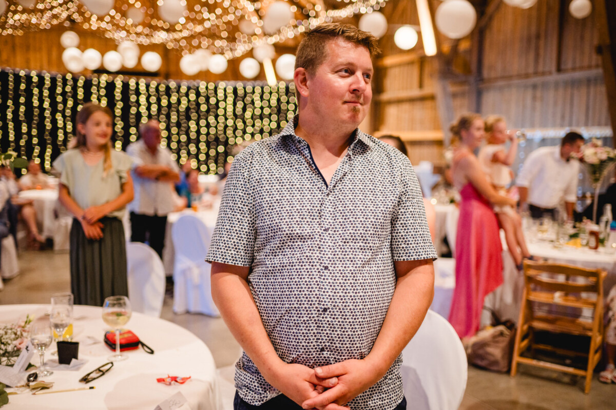 Ein Mann im gemusterten Hemd steht in einem dekorierten Saal mit Lichterketten und Tischen, im Hintergrund haben sich Menschen versammelt.