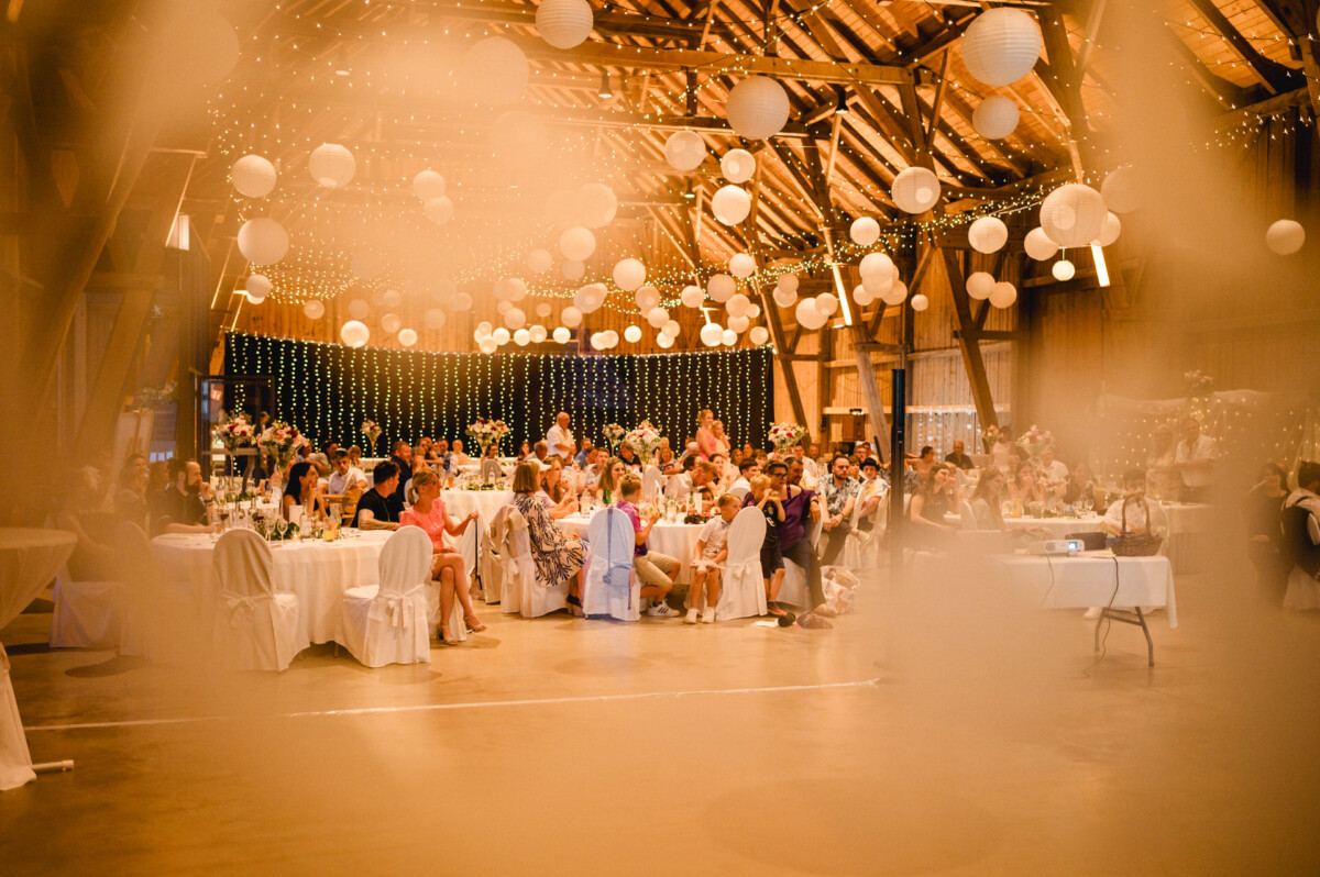 Eine große Indoor-Veranstaltung mit Menschen, die an runden Tischen sitzen. Die Decke ist mit Lichterketten und Papierlaternen dekoriert.