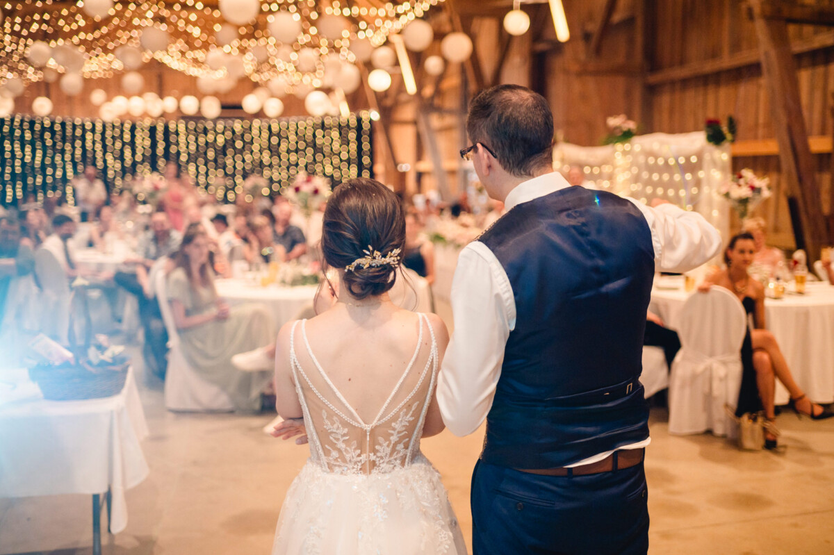Ein Paar in Abendgarderobe steht in einem mit Lichterketten dekorierten Veranstaltungsort vor einem sitzenden Publikum.