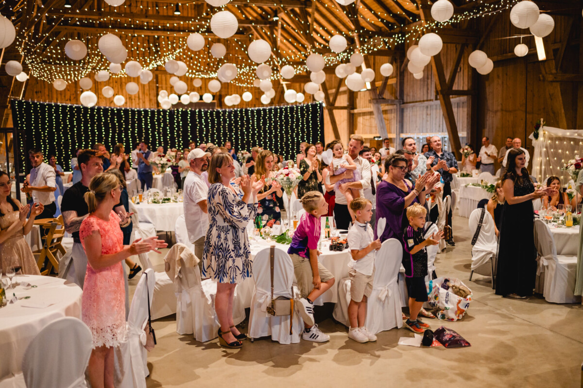 Eine Gruppe von Menschen in Abendgarderobe steht in einer mit Lichterketten und Papierlaternen dekorierten Scheune und applaudiert.