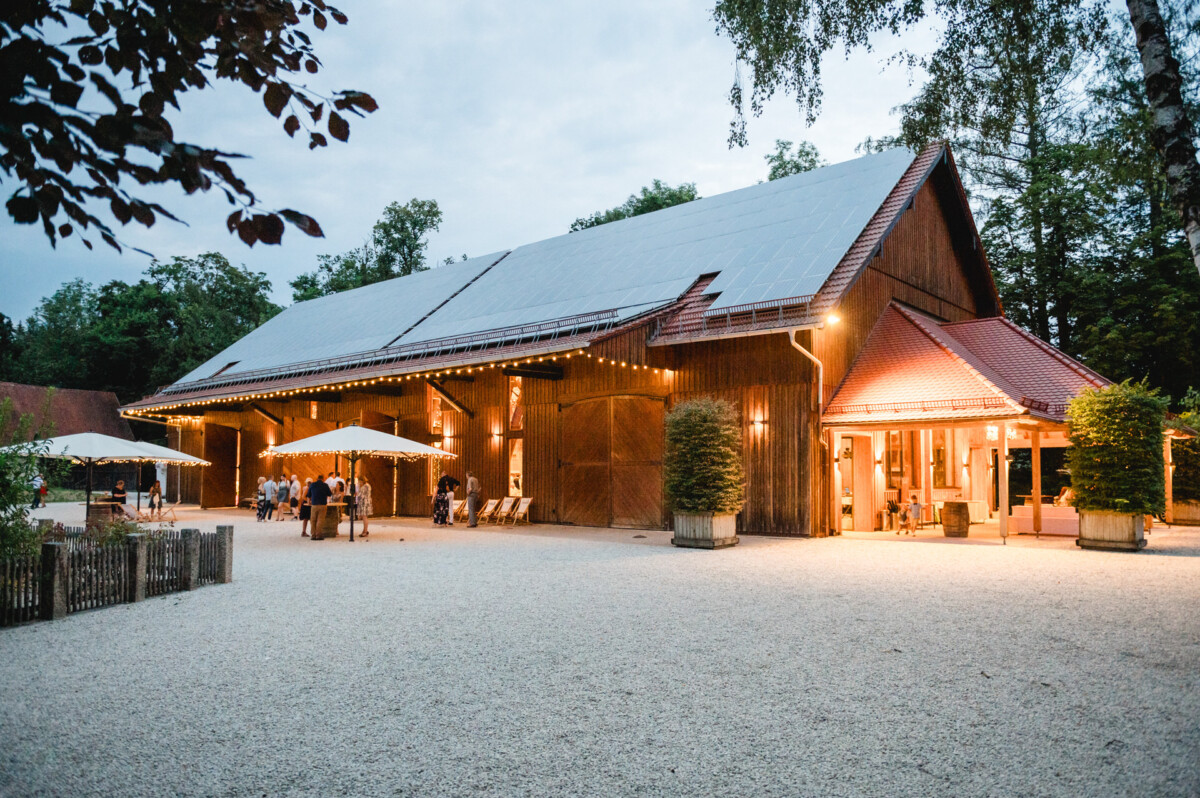 Eine große Holzscheune mit modernem Dach, umgeben von Bäumen. Die Scheune ist mit Lichterketten beleuchtet und draußen auf einer Kiesfläche sitzen Menschen an Tischen mit Sonnenschirmen.