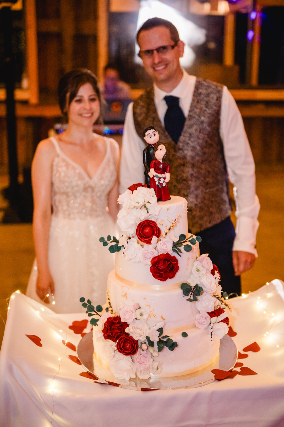 Braut und Bräutigam stehen hinter einer dreistöckigen Hochzeitstorte, die mit roten und weißen Blumen und einer Figur als Aufsatz geschmückt ist, auf einem Tisch mit verstreuten Rosenblättern.