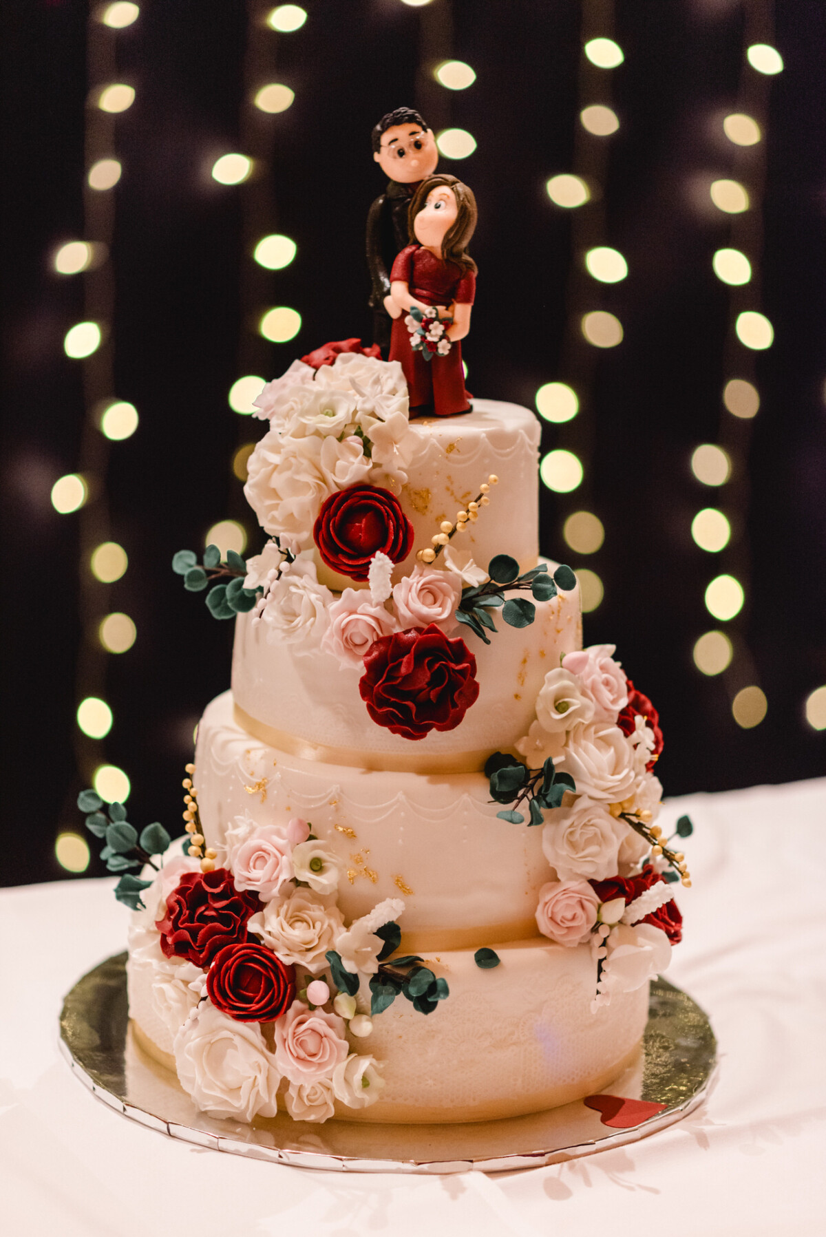 Eine dreistöckige Hochzeitstorte mit roten und weißen Rosen und einer Paarfigur als Krönung vor einem Hintergrund aus verschwommenen Lichtern.