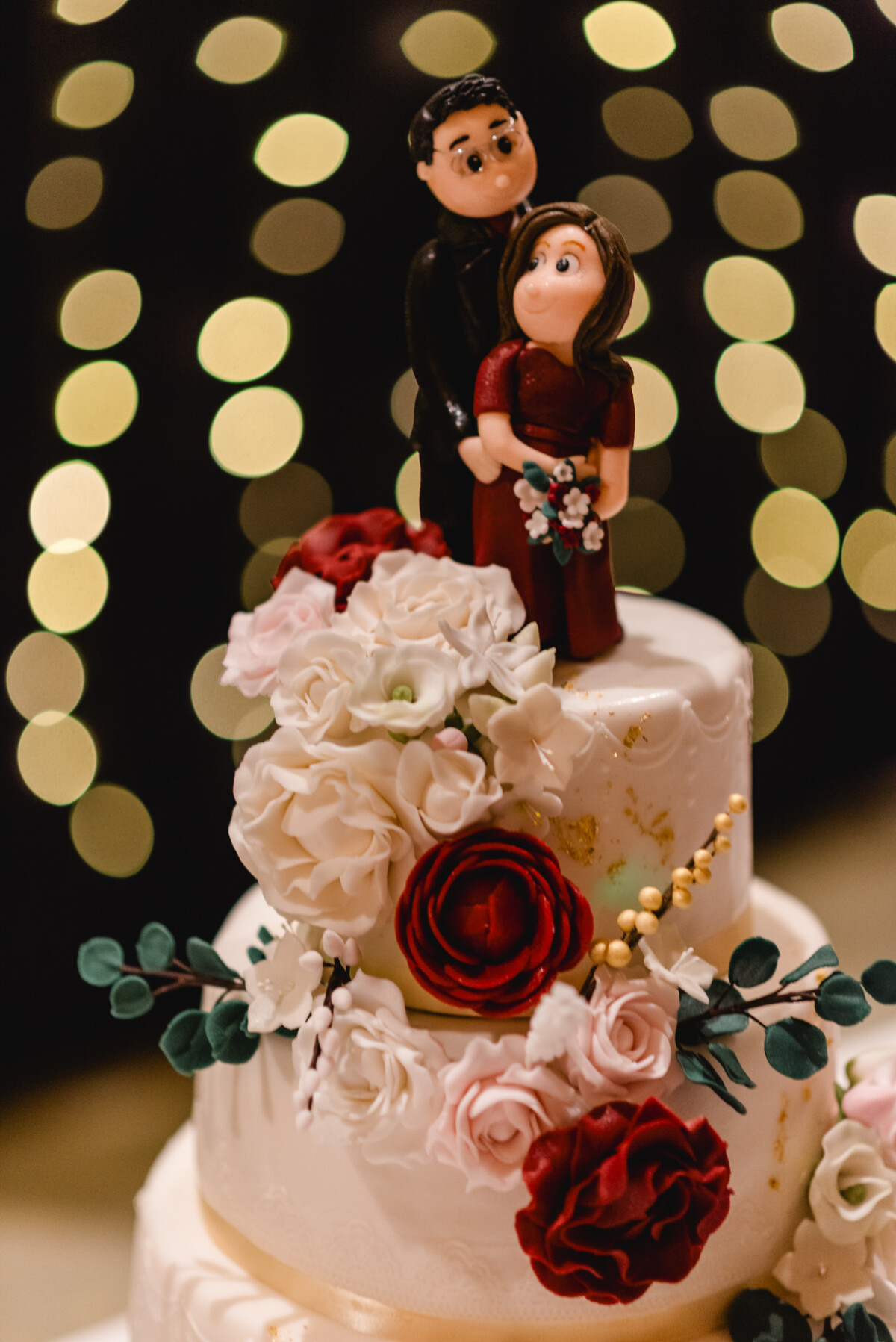 Eine Hochzeitstorte, gekrönt von einem Paar in Form einer Figur, umgeben von weißen und roten Blumen, vor einem Hintergrund aus verschwommenen gelben Lichtern.