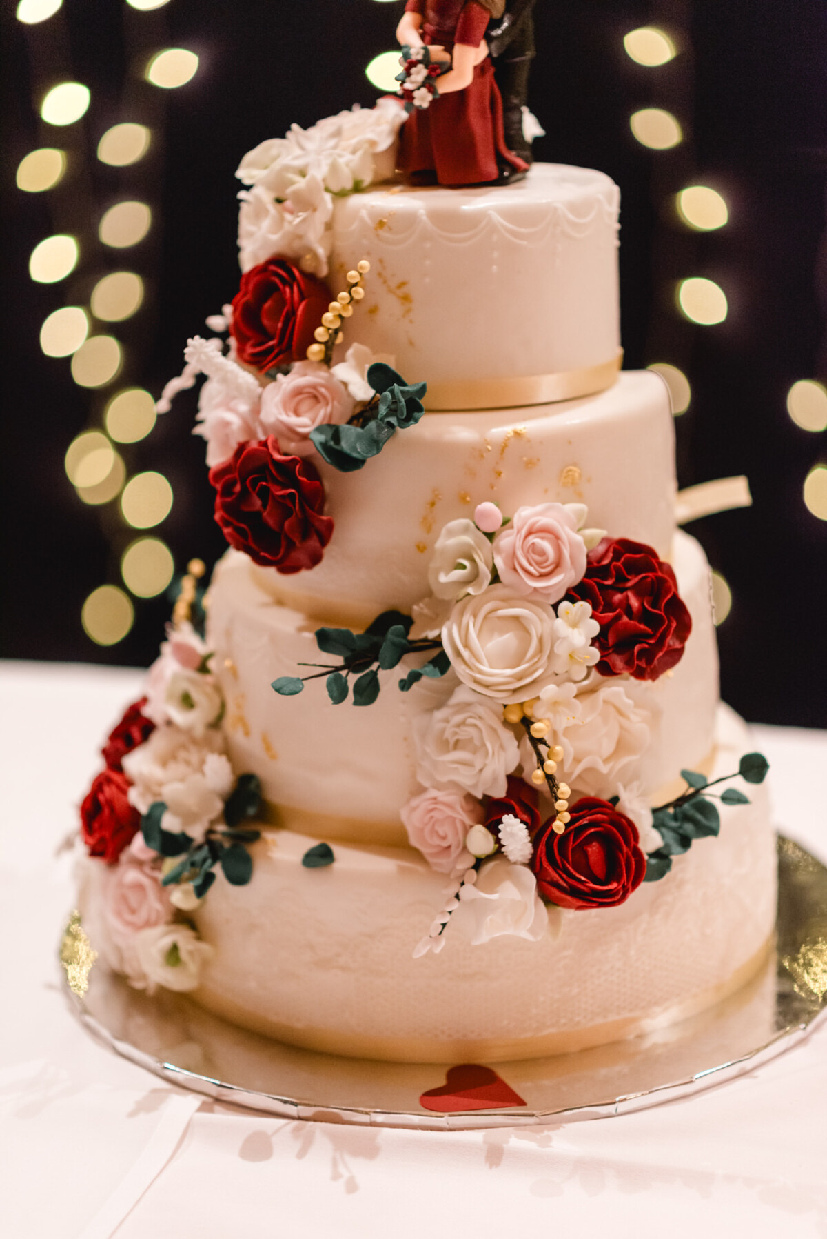 Eine dreistöckige weiße Hochzeitstorte, geschmückt mit roten und weißen Blumen, Beeren und Blättern vor einem Hintergrund aus verschwommenen Lichtern.
