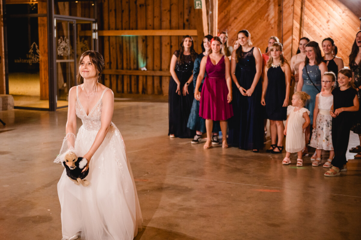 Eine Braut in einem weißen Kleid hält ein kleines Stofftier und bereitet sich darauf vor, es einer Gruppe gespannt wartender Frauen und Kinder in einer Scheune zuzuwerfen.