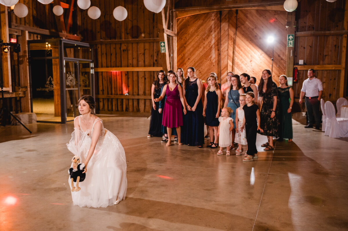 Eine Braut ist im Begriff, ein Stofftier über ihre Schulter zu einer Gruppe von Frauen und Kindern zu werfen, die in einer Reihe hinter ihr in einem rustikalen Holzsaal stehen.