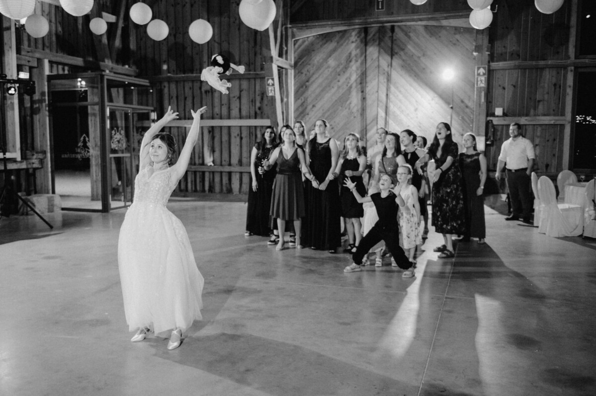 Die Braut wirft einer Gruppe von Frauen und Kindern in einer Scheune einen Blumenstrauß zu, von deren Decke dekorative Laternen hängen.