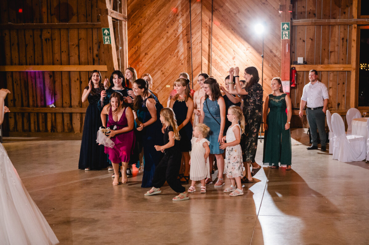 Eine Gruppe von Frauen und Kindern steht aufgeregt in einer scheuneähnlichen Anlage und bereitet sich darauf vor, einen Blumenstrauß zu fangen.