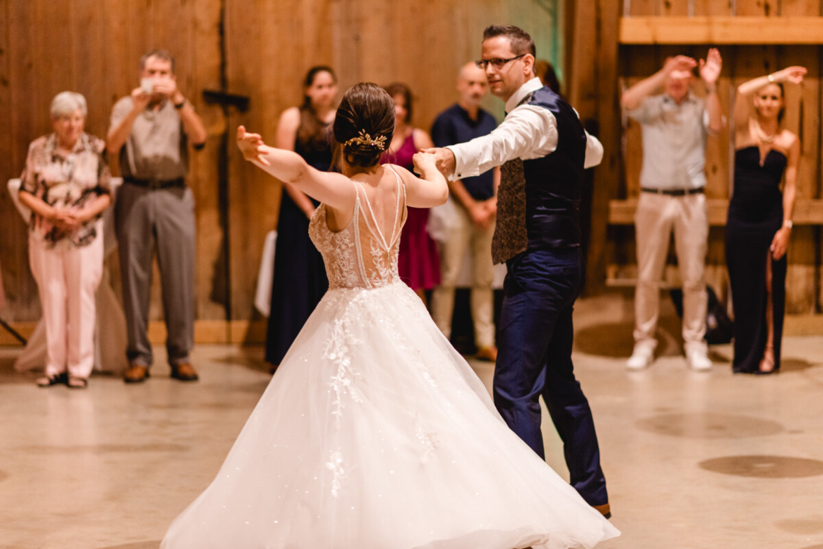 Ein Paar tanzt in einer Scheune, sie trägt ein weißes Kleid mit vielen Details und er ist in Abendgarderobe. Im Hintergrund schauen die Gäste zu, manche halten den Moment mit ihren Handys fest.