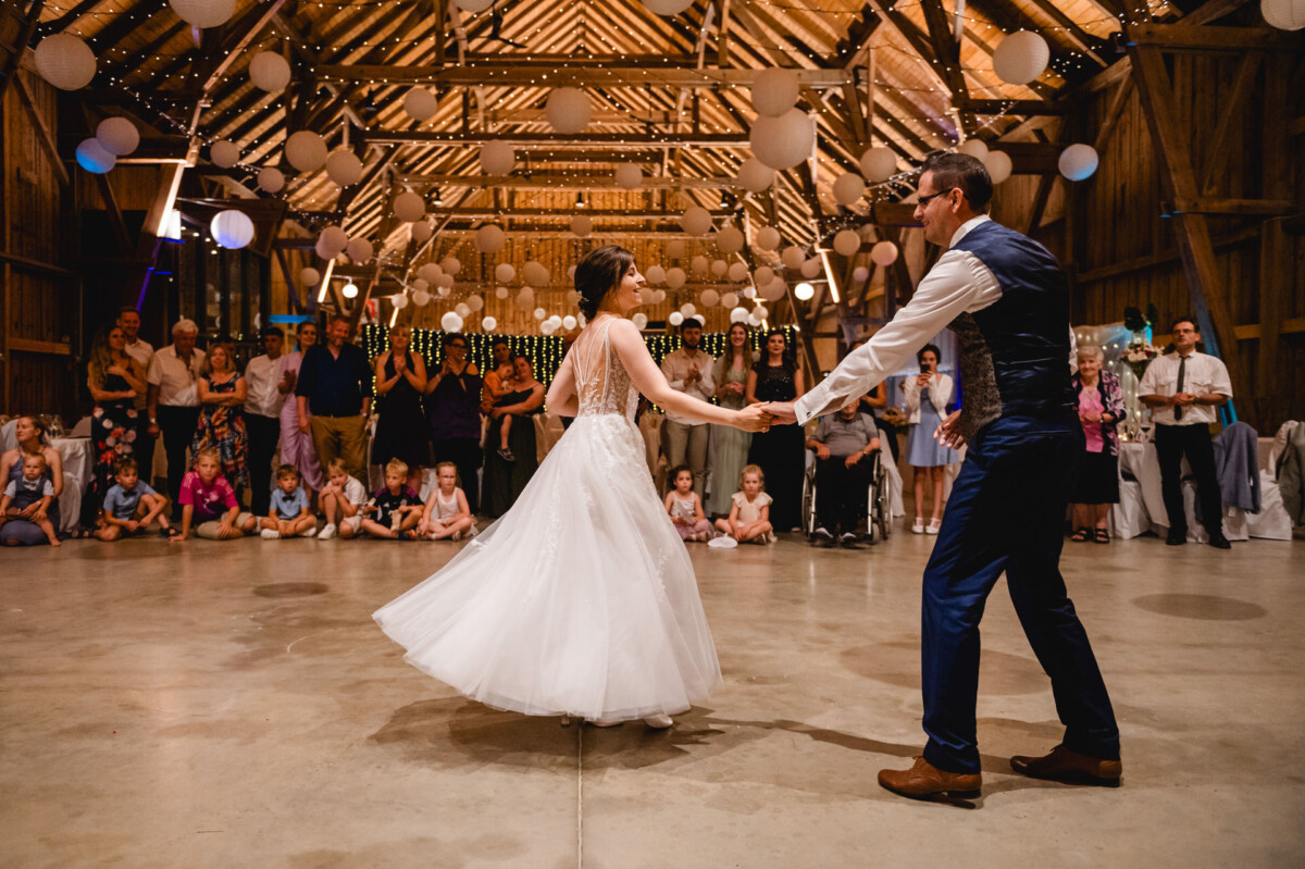 Braut und Bräutigam tanzen in einer mit Hängelampen und Laternen dekorierten Scheune, umgeben von sitzenden und stehenden Gästen.