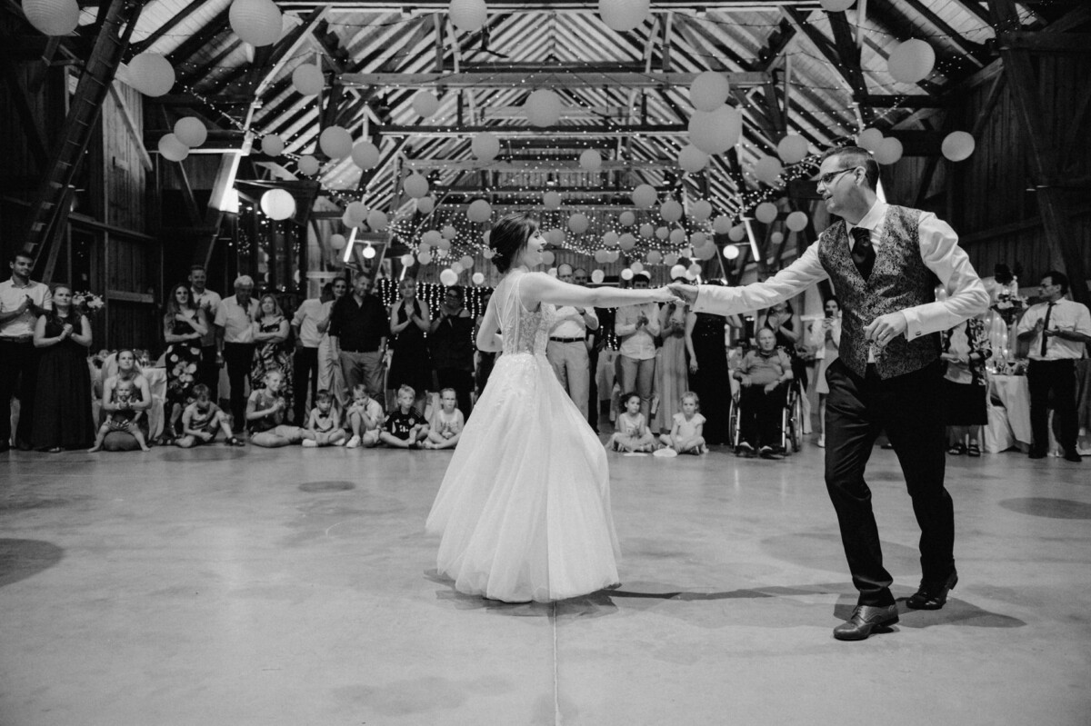 Braut und Bräutigam tanzen in einer dekorierten Scheune, während die Gäste sitzend und stehend zuschauen.