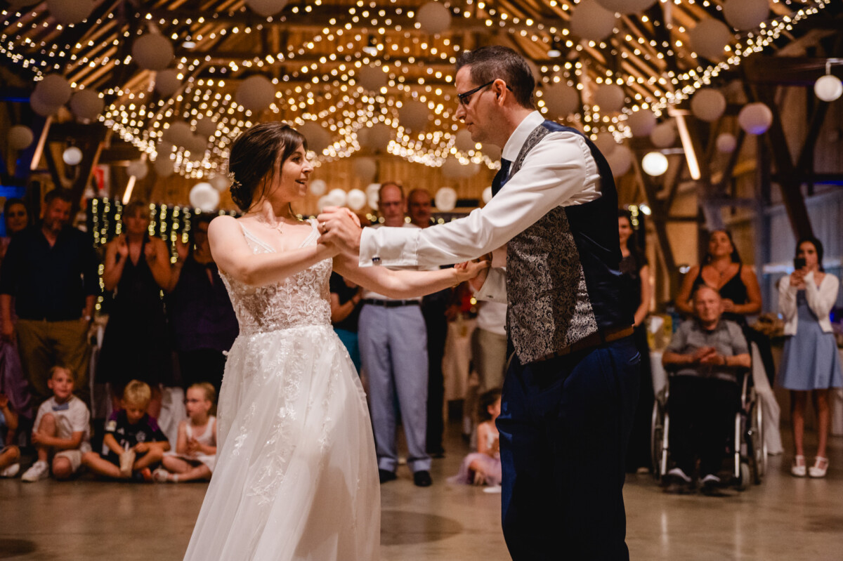 Ein Paar tanzt bei einer Hochzeitsfeier unter Lichterketten, umgeben von Gästen, darunter Kinder und eine Person im Rollstuhl.