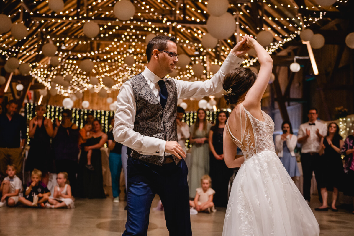 Ein Paar tanzt in einem warm beleuchteten, mit Lichterketten geschmückten Veranstaltungsort, umgeben von einem Publikum aus sitzenden und stehenden Gästen.