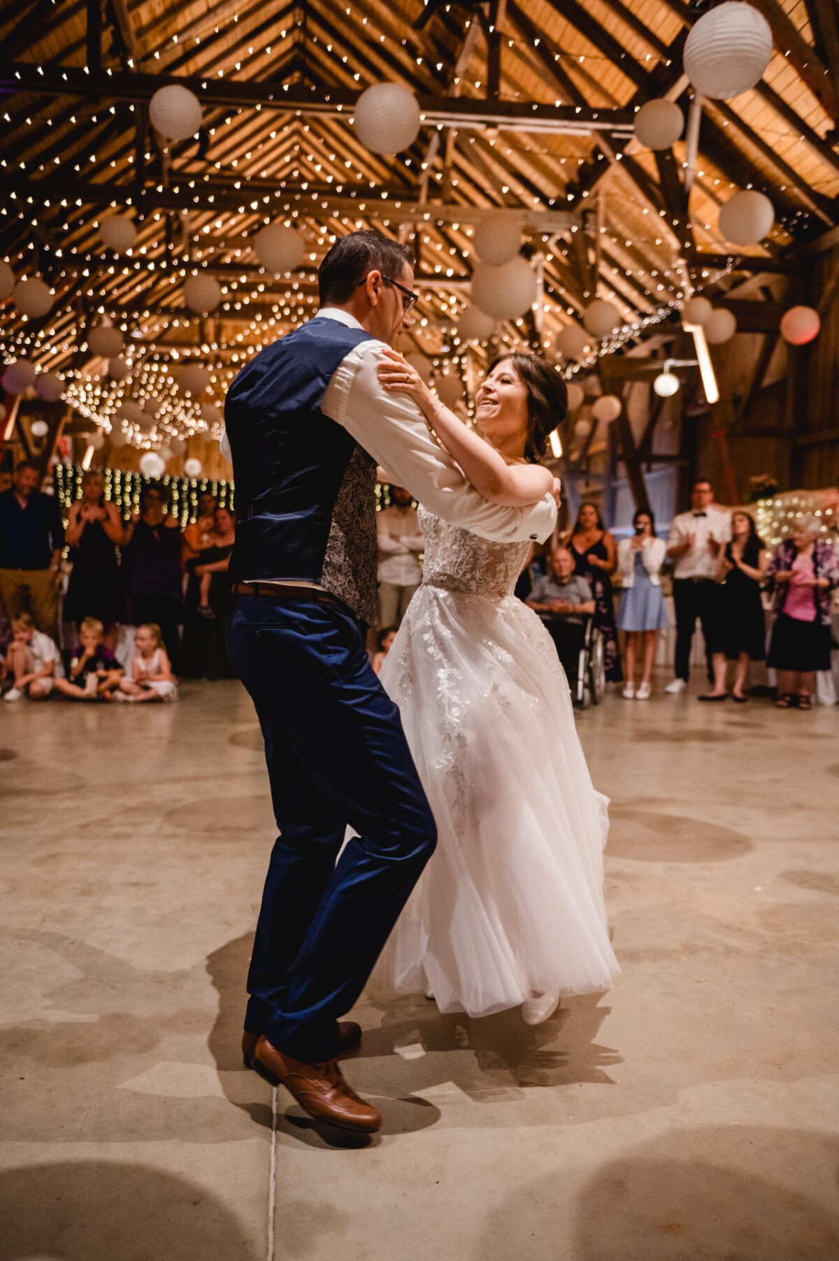 Ein Paar in Abendgarderobe tanzt fröhlich auf dem dekorierten Boden eines Ballsaals, umgeben von einem Publikum und funkelnden Lichtern.
