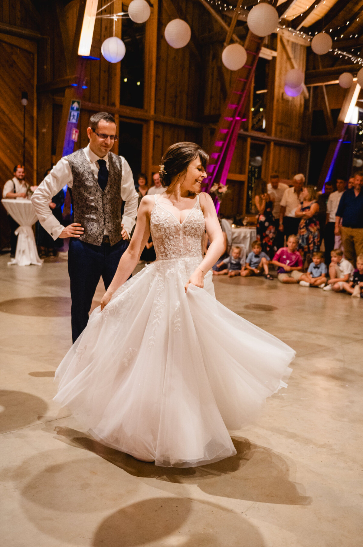 Braut und Bräutigam tanzen umgeben von Gästen in einem rustikalen Veranstaltungsort, der mit Hängelampen und weißen Papierlaternen dekoriert ist.