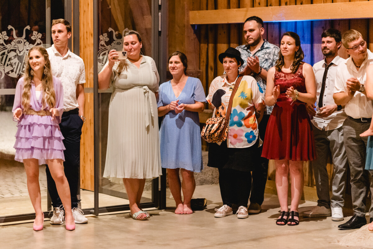 Eine Gruppe von Menschen steht drinnen, einige klatschen und andere halten Getränke in der Hand. Mit einem hölzernen Interieur im Hintergrund scheint es sich um eine zwanglose Veranstaltung zu handeln.