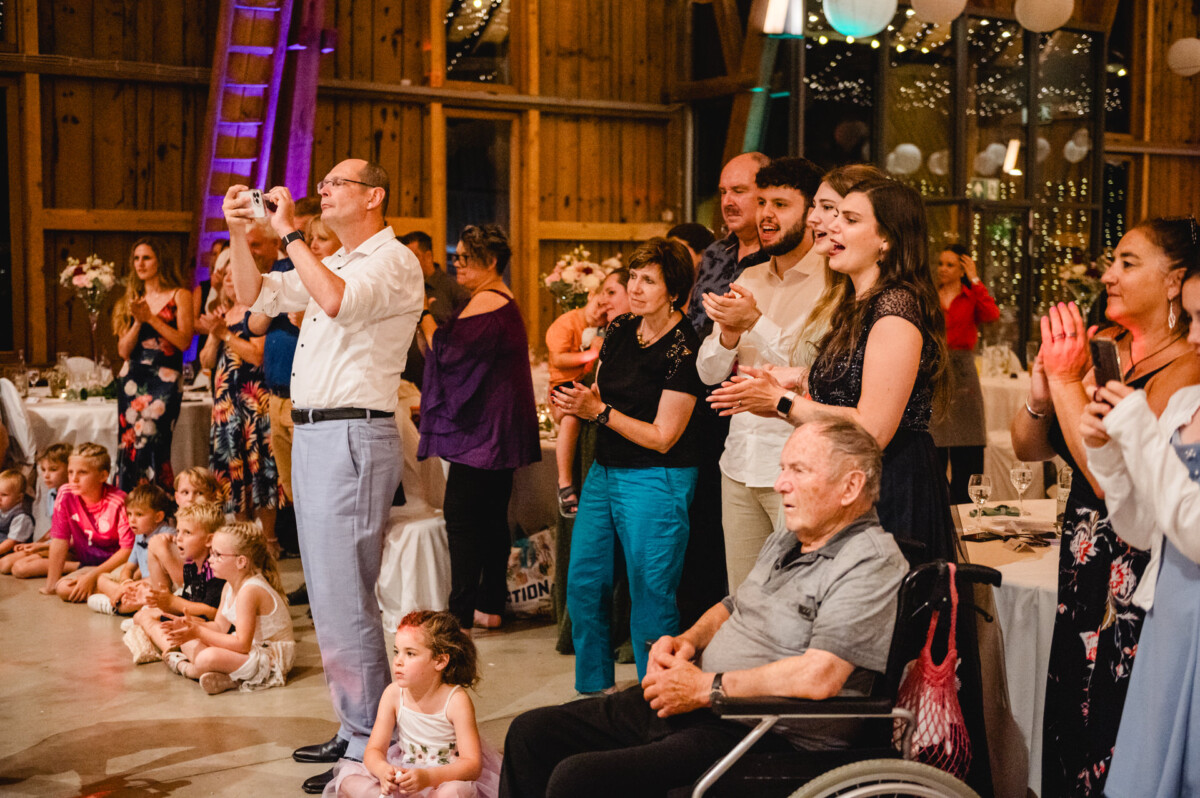 Eine Gruppe von Menschen, darunter Kinder und ein Mann im Rollstuhl, beobachtet außerhalb des Kamerablickfelds eine Veranstaltung in einer mit Lichtern und Blumen geschmückten Holzhalle.