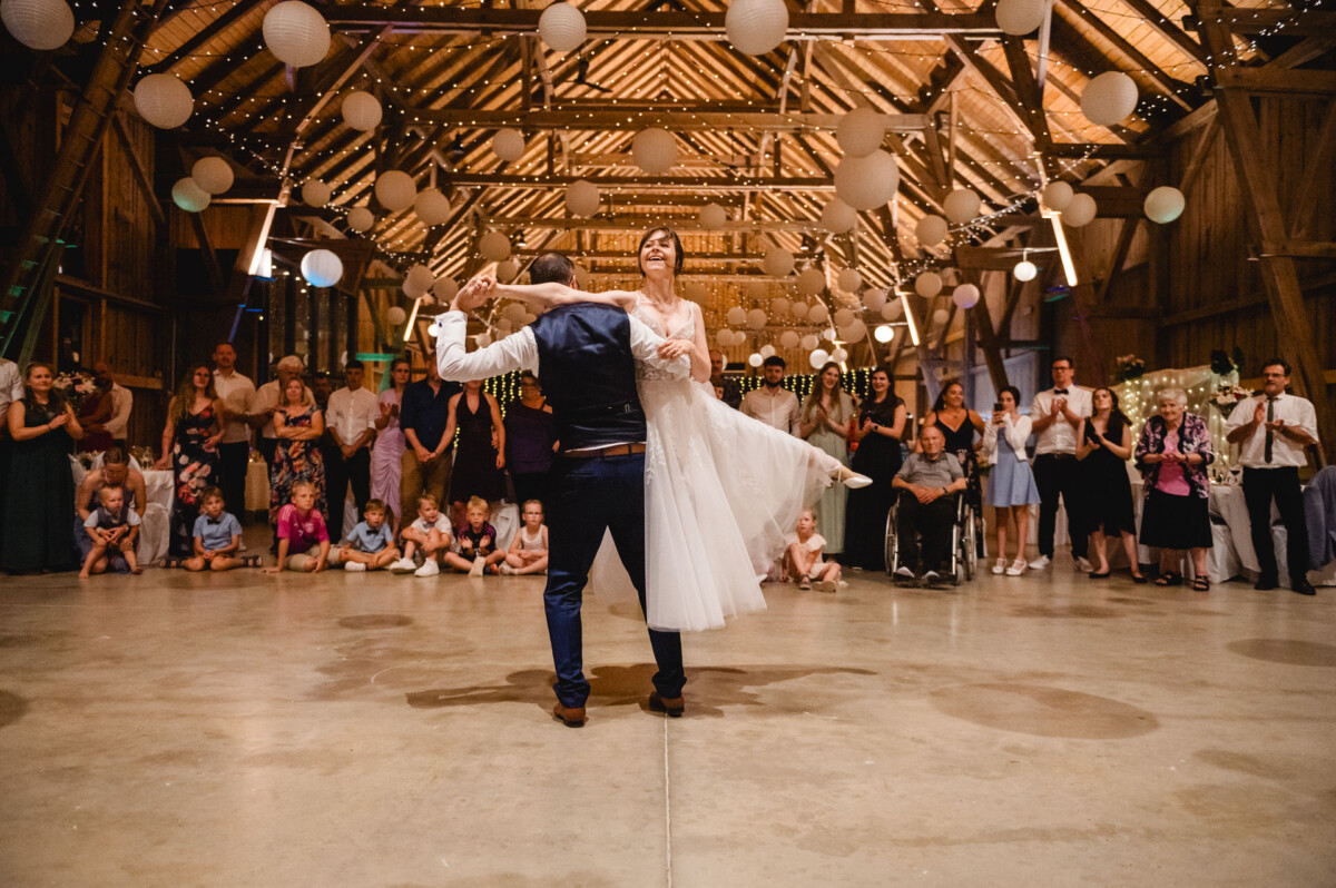 Ein Paar tanzt während einer Hochzeitsfeier in der Mitte einer rustikalen Scheune, umgeben von sitzenden und stehenden Gästen. Laternen und Lichterketten schmücken die Decke.