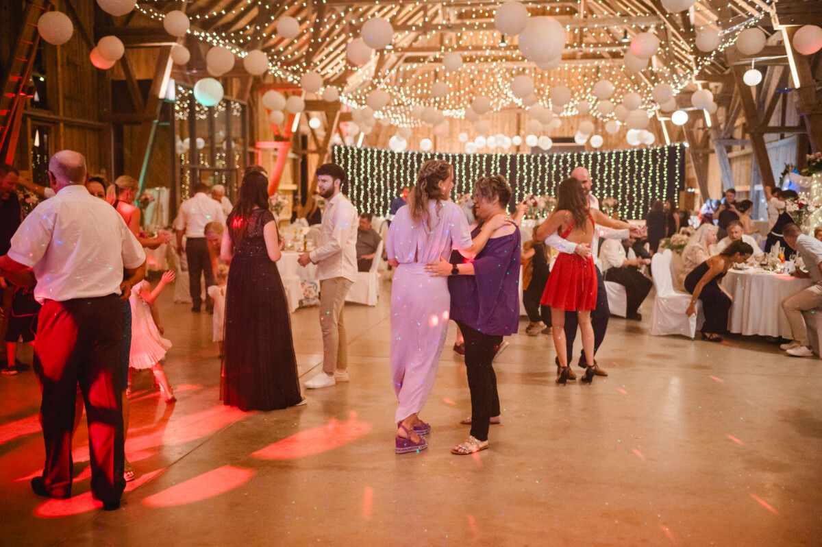 Menschen tanzen in einer mit Lichterketten und hängenden Papierlaternen dekorierten Scheune. Im Hintergrund sitzen Gäste an Tischen und die Tanzfläche wird von roten Scheinwerfern beleuchtet.