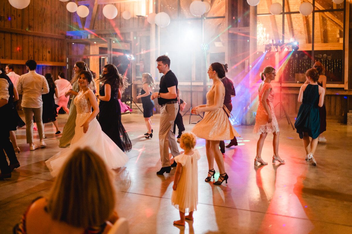In einem warm beleuchteten, rustikalen Saal mit Holzwänden und bunten Lichtern wird getanzt. Im Vordergrund steht ein Kind und beobachtet die Tänzer.