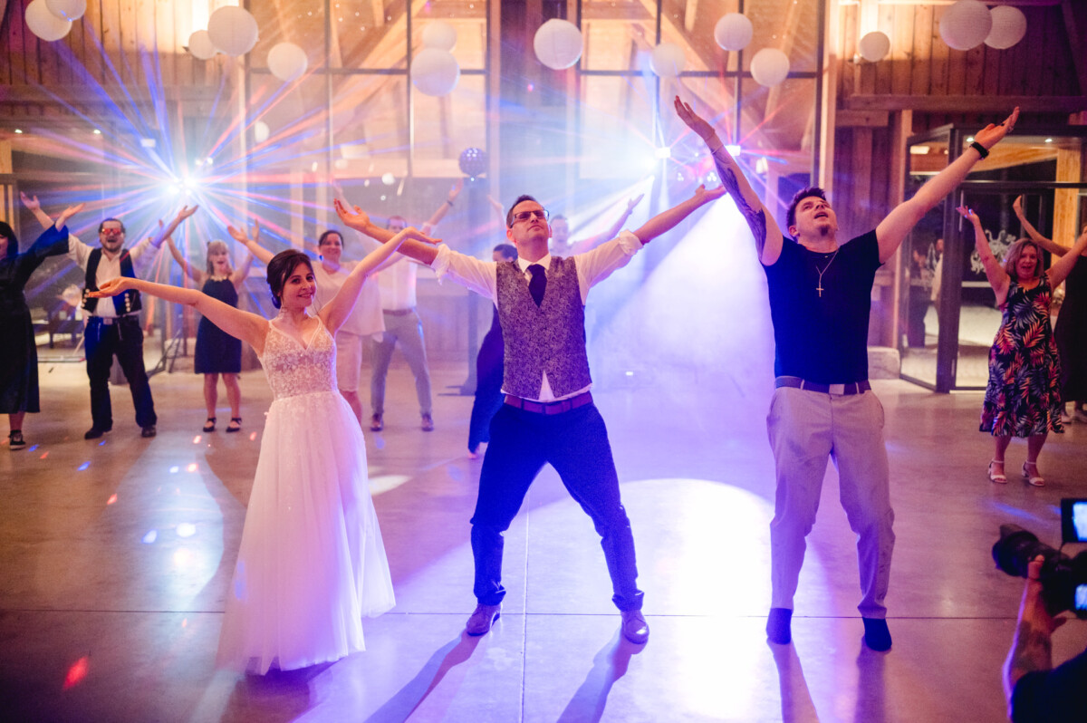 Menschen, darunter eine Braut und ein Bräutigam, tanzen in einer Scheune mit bunten Lichtern und Dekorationen.