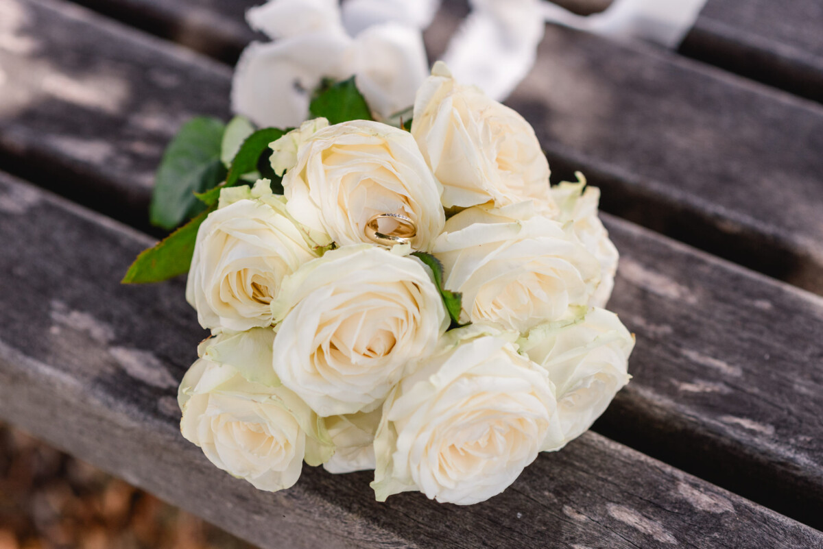 Ein Strauß weißer Rosen mit zwei dazwischen platzierten goldenen Ringen, ruht auf einer verwitterten Holzoberfläche.