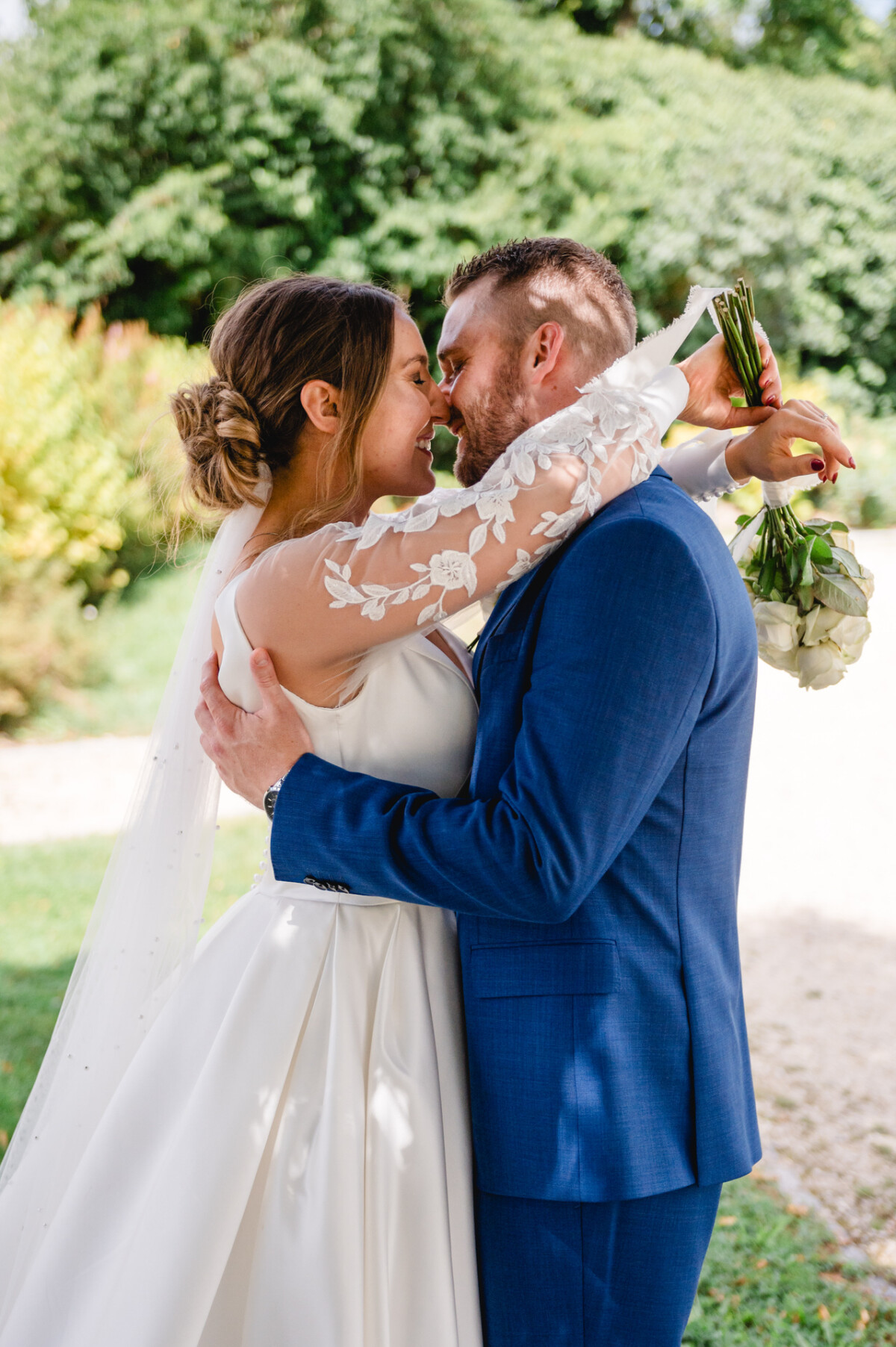 Braut und Bräutigam umarmen und küssen sich im Freien, wobei die Braut einen Blumenstrauß hält. Im Hintergrund sind Grünflächen und ein Kiesweg zu sehen.