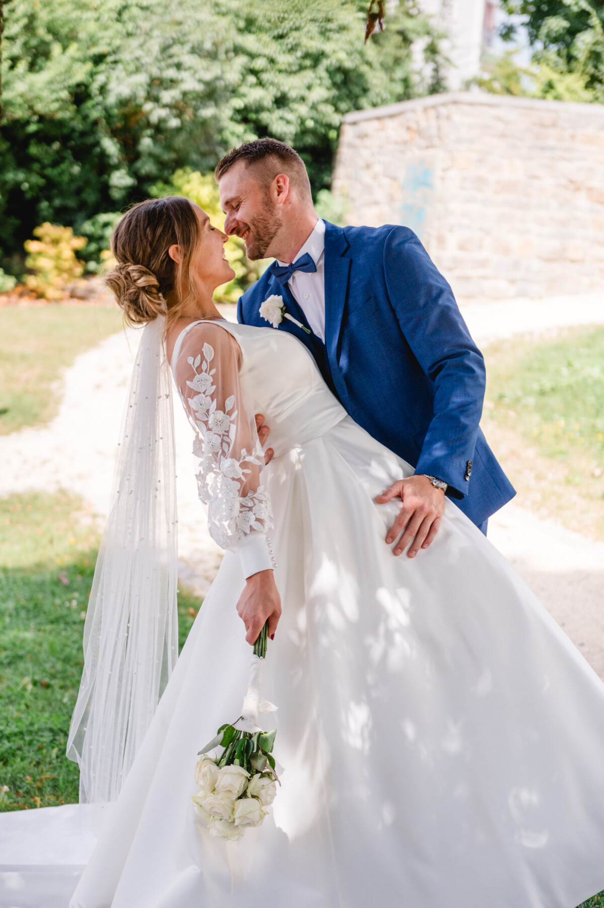 Ein Paar posiert für ein Hochzeitsfoto im Freien, wobei der Bräutigam die Braut ins Gesicht taucht. Sie hält einen Strauß weißer Rosen und beide tragen formelle Hochzeitskleidung.